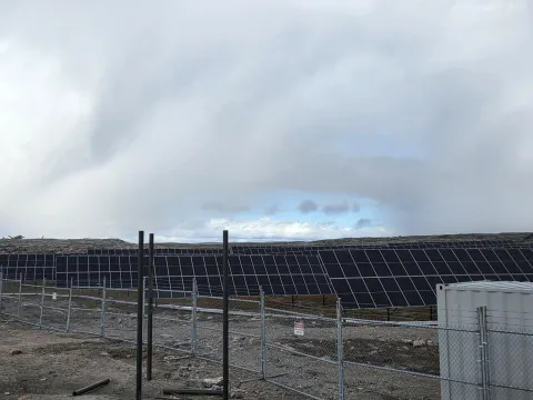 Rows of solar panels installed on rocky tundra behind a chain-link fence under an overcast sky, with a small equipment enclosure in the foreground.