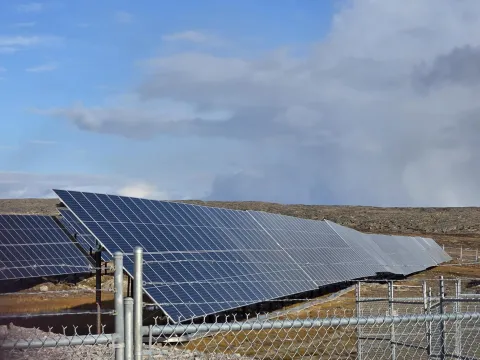 Rows of solar panels installed on rocky tundra behind a chain-link fence under an overcast sky, with a small equipment enclosure in the foreground.
